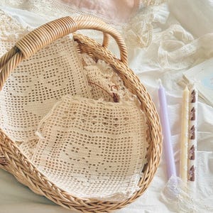 May include: A round woven wicker basket with a handle containing several off-white crocheted squares with intricate patterns. Beside the basket are two slender taper candles, one solid light purple and one cream with a brown rabbit print.
