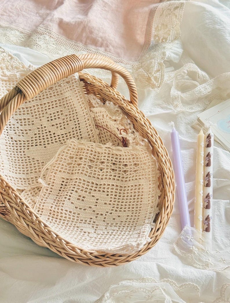 May include: A round woven wicker basket with a handle containing several off-white crocheted squares with intricate patterns. Beside the basket are two slender taper candles, one solid light purple and one cream with a brown rabbit print.