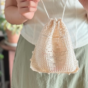 May include: A cream-colored drawstring pouch crafted with an open-work crochet pattern. The bag features a square base, a grid-like texture throughout the body, and a delicate scalloped edge along the bottom. It is secured with two thin cords.