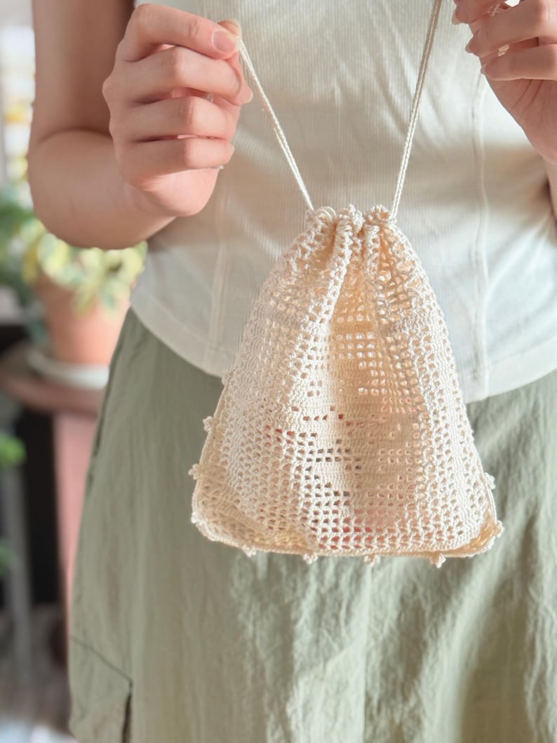 May include: A cream-colored drawstring pouch crafted with an open-work crochet pattern. The bag features a square base, a grid-like texture throughout the body, and a delicate scalloped edge along the bottom. It is secured with two thin cords.