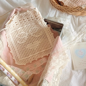 May include: A cream-colored square crochet doily with a central heart motif, featuring a scalloped edge. The doily rests on a pink fabric lining inside a metal container, accompanied by a small card labeled vanilla crochet with a heart graphic.
