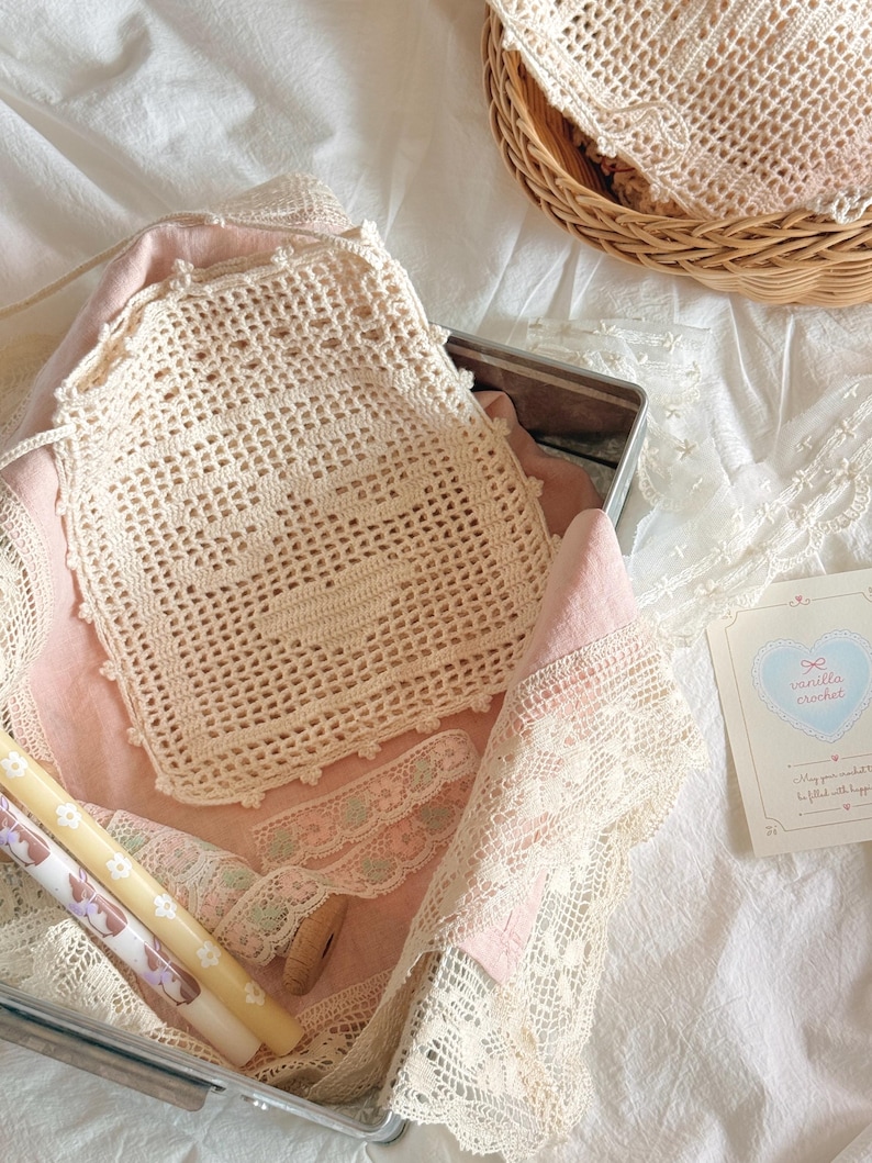 May include: A cream-colored square crochet doily with a central heart motif, featuring a scalloped edge. The doily rests on a pink fabric lining inside a metal container, accompanied by a small card labeled vanilla crochet with a heart graphic.