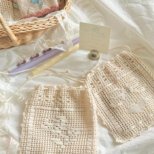 May include: Two off-white crochet drawstring pouches featuring a heart pattern, a wicker basket containing a small floral crochet item, two taper candles with rabbit and flower prints, and a small card with text held by a metal stand.