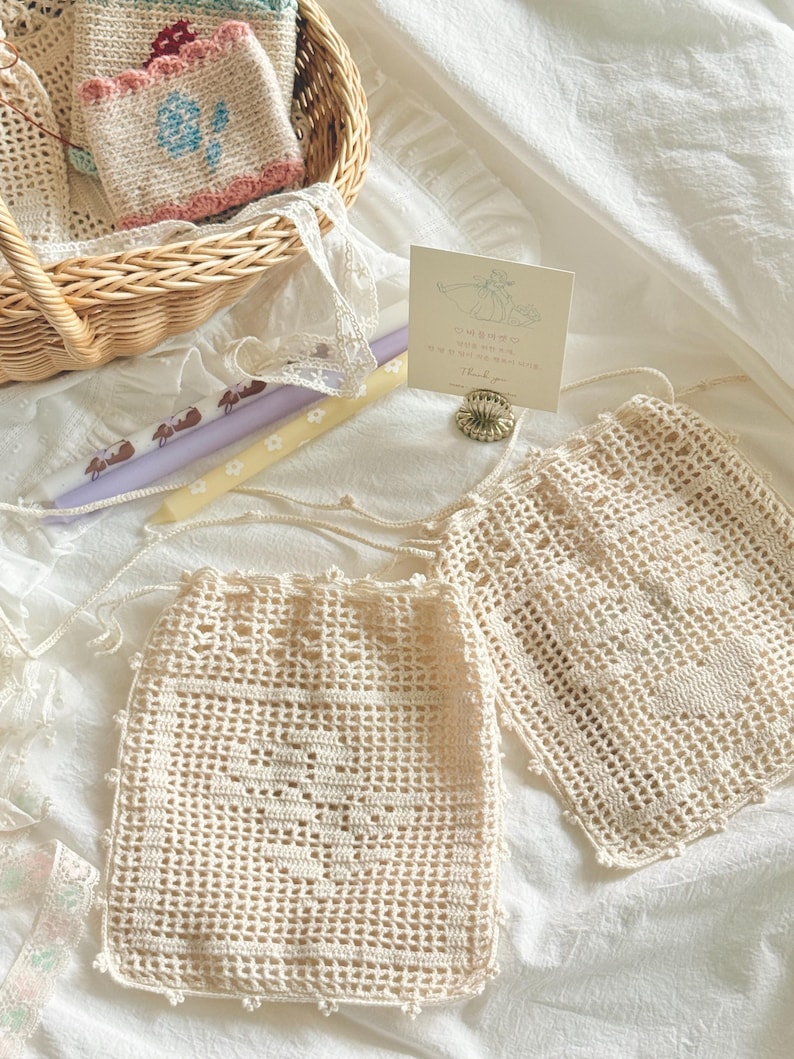May include: Two off-white crochet drawstring pouches featuring a heart pattern, a wicker basket containing a small floral crochet item, two taper candles with rabbit and flower prints, and a small card with text held by a metal stand.