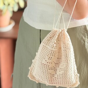 May include: A cream-colored drawstring pouch crafted from intricate crochet lace. The bag features a delicate openwork grid pattern and a scalloped edge along the bottom. It is equipped with two thin, matching cream-colored cord handles for carrying.