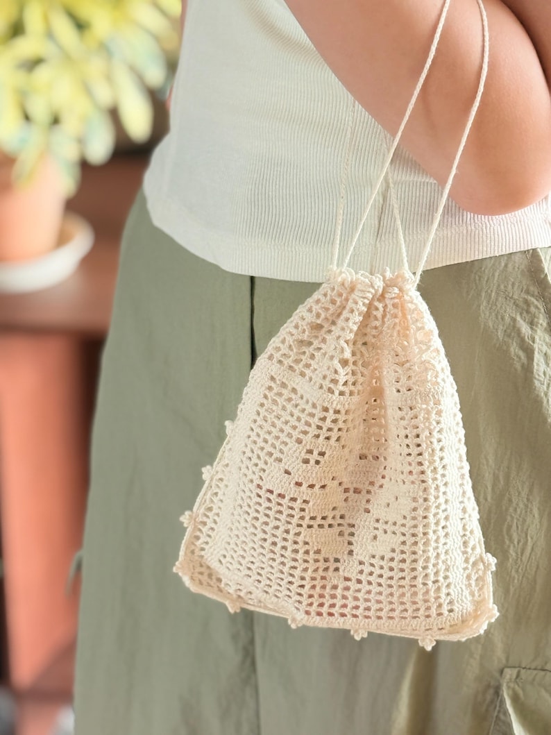 May include: A cream-colored drawstring pouch crafted from intricate crochet lace. The bag features a delicate openwork grid pattern and a scalloped edge along the bottom. It is equipped with two thin, matching cream-colored cord handles for carrying.