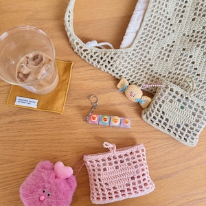 Op de afbeelding: Een verzameling accessoires op een houten oppervlak. Inclusief een glas met een drankje, een gele onderzetter met tekst, een gehaakte tote bag, een roze katvormig pluche met een hart, een roze gehaakt zakje en een sleutelhanger met snoep.