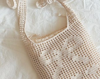 May include: A cream-colored, open-weave crochet tote bag with a long strap and a decorative bow detail at the top. The bag is made of a soft, natural fiber and has a rectangular shape.