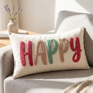 Könnte beinhalten: Cremefarbenes rechteckiges Kissen mit dem Wort "HAPPY" in strukturierten Buchstaben gestickt. Die Buchstaben sind in Rot-, Beige- und Grüntönen gehalten. Das Kissen liegt auf einem grauen Sofa mit einer beigen Decke.
