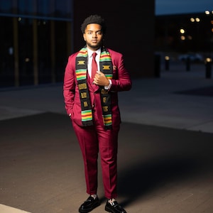 May include: A person wearing a burgundy suit, white shirt, and red tie, with a graduation stole. The stole is black with gold and green accents and text that reads "Educated Black Men Class of 2023". Black dress shoes complete the outfit.