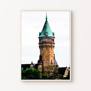 Può includere: Stampa incorniciata di un'alta torre dell'orologio con un tetto in rame verde e un quadrante dorato. La torre è in pietra beige ed è circondata da alberi verdi e altri edifici. Il cielo è coperto.