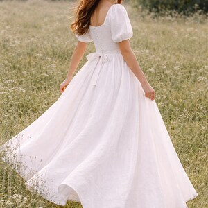 May include: A white linen dress with a square neckline, short puff sleeves, and a flowing skirt. The dress features a fitted bodice with a bow at the waist. The model is wearing brown sandals and is in a field of wildflowers.