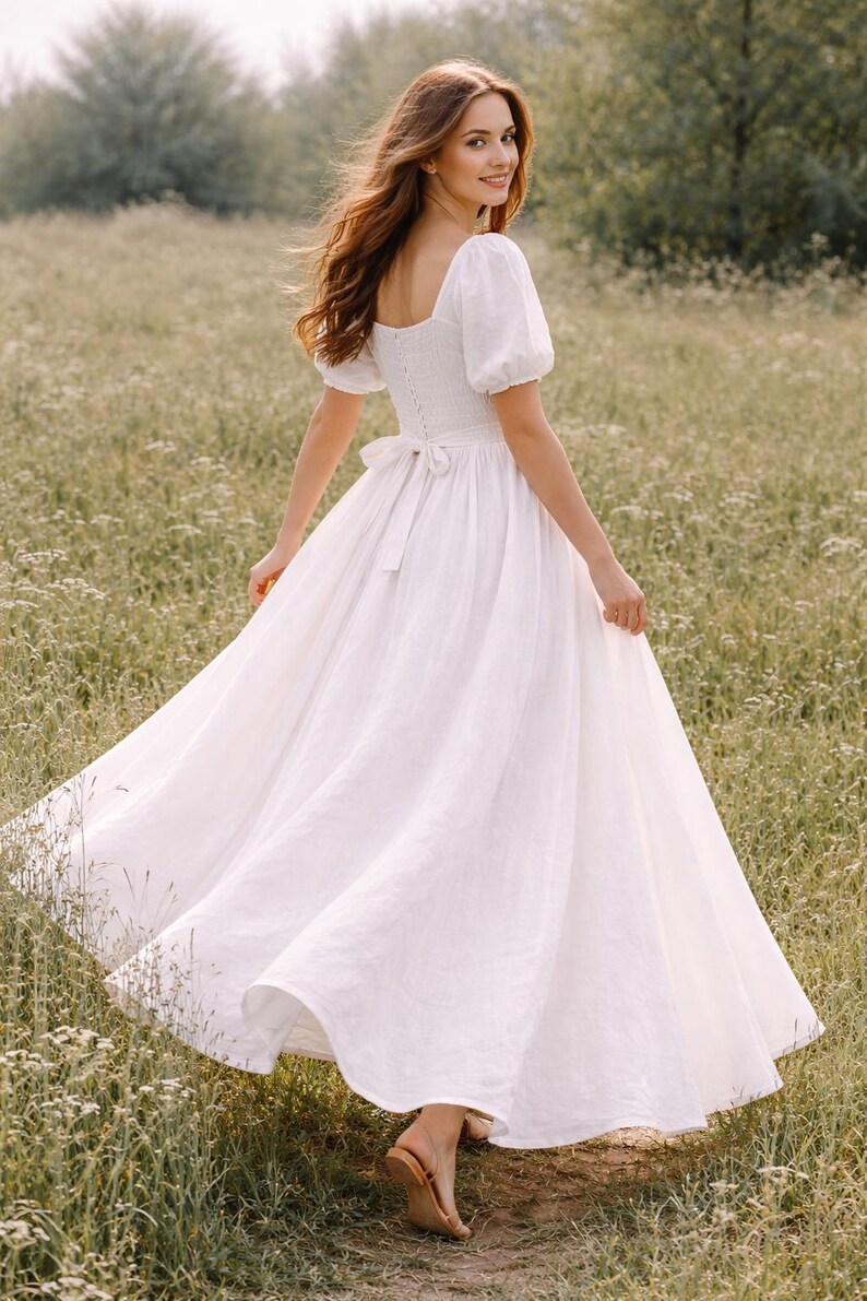May include: A white linen dress with a square neckline, short puff sleeves, and a flowing skirt. The dress features a fitted bodice with a bow at the waist. The model is wearing brown sandals and is in a field of wildflowers.