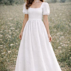 May include: A white linen dress with a square neckline and short, puffed sleeves. The dress has a fitted bodice and a flowing, floor-length skirt. The model is standing in a field of wildflowers.