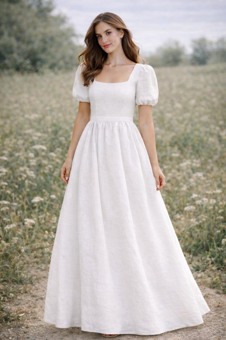 May include: A white linen dress with a square neckline and short, puffed sleeves. The dress has a fitted bodice and a flowing, floor-length skirt. The model is standing in a field of wildflowers.