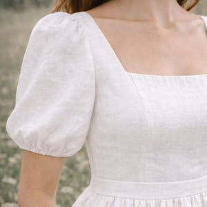 May include: A white linen dress with a square neckline and short, puffed sleeves. The dress features a fitted bodice and a gathered skirt, creating a classic and elegant silhouette. The fabric appears to be lightweight and breathable, suitable for warm weather.