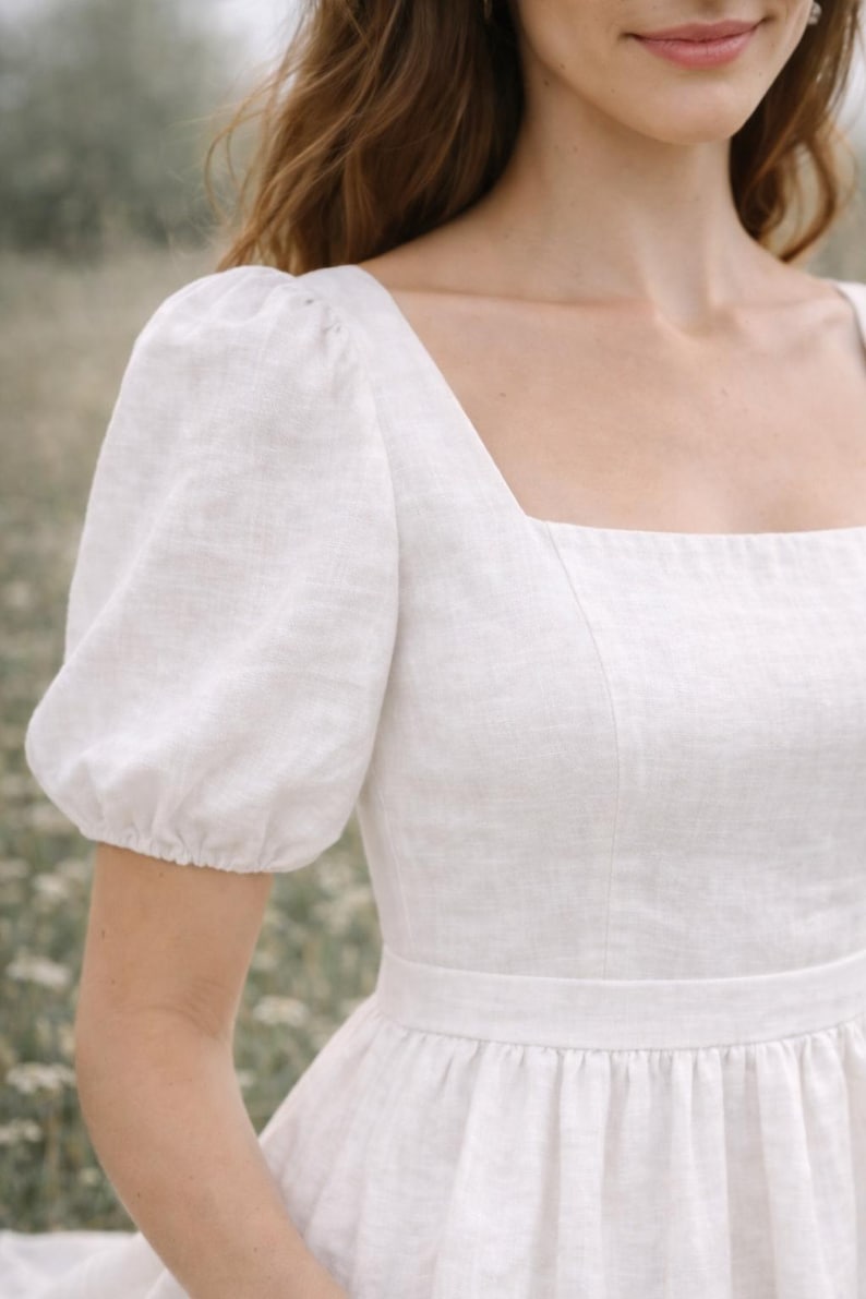 May include: A white linen dress with a square neckline and short, puffed sleeves. The dress features a fitted bodice and a gathered skirt, creating a classic and elegant silhouette. The fabric appears to be lightweight and breathable, suitable for warm weather.