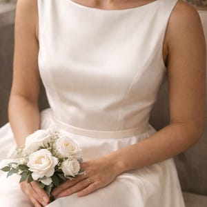 May include: An ivory wedding dress with a boat neck and a fitted bodice. The bride holds a small bouquet of white roses and greenery. The dress is sleeveless and has a simple, elegant design, suitable for a wedding ceremony.