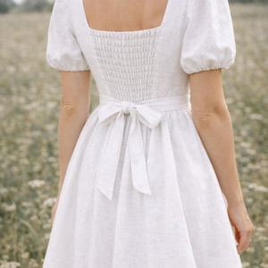 May include: White linen dress with a square neckline, short puff sleeves, and a fitted bodice with a smocked back. The dress features a waist tie and a flowing skirt, perfect for a summer day.