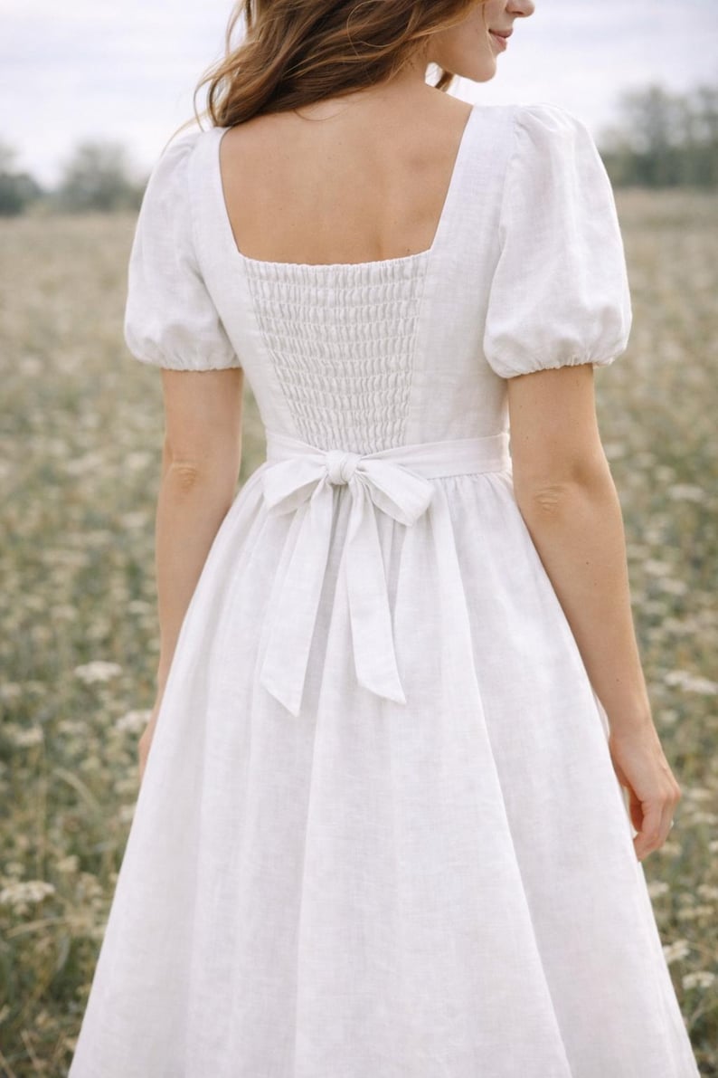 May include: White linen dress with a square neckline, short puff sleeves, and a fitted bodice with a smocked back. The dress features a waist tie and a flowing skirt, perfect for a summer day.