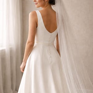 May include: A white wedding dress with a low scoop back and a row of buttons down the centre. The dress has a simple, elegant design with a flowing skirt. A sheer veil drapes over the back of the dress.