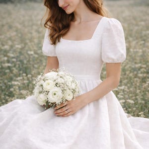 May include: A white linen wedding dress with a square neckline and short, puffed sleeves. The bride holds a bouquet of white flowers. The dress has a fitted bodice and a flowing skirt, perfect for a garden wedding.