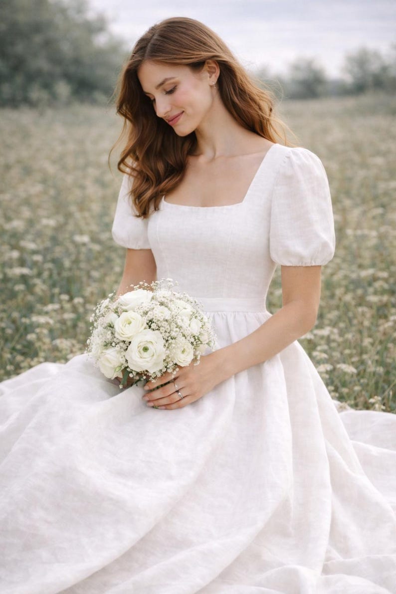 May include: A white linen wedding dress with a square neckline and short, puffed sleeves. The bride holds a bouquet of white flowers. The dress has a fitted bodice and a flowing skirt, perfect for a garden wedding.
