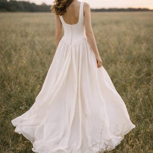 May include: A white wedding dress with a square neckline and a flowing skirt. The dress has a fitted bodice with small buttons down the back. The model is standing in a field of tall grass.