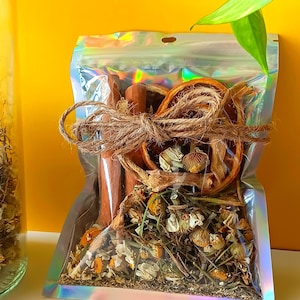 May include: A clear, iridescent pouch filled with dried herbs, spices, and a dried orange slice, tied with twine. Cinnamon sticks and dried flowers are visible. The background is a bright yellow.