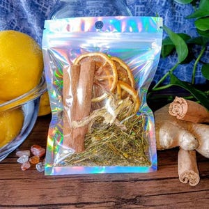 Puede incluir: Un frasco de vidrio transparente con tapa dorada está detrás de una bolsa holográfica con cremallera llena de rodajas de naranja secas, palitos de canela y hierbas. También se ve un cuenco de limones y raíces de jengibre. Los artículos están dispuestos sobre una superficie de madera.