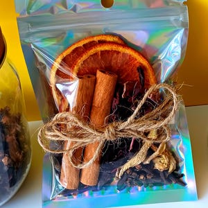 May include: A clear, iridescent pouch filled with dried orange slices, cinnamon sticks, and various spices. The pouch is tied with twine and sits against a bright yellow background. The contents appear to be for a tea or potpourri blend.
