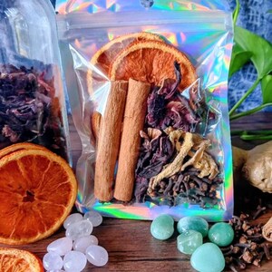 May include: A clear glass jar and a holographic bag filled with dried herbs, orange slices, and cinnamon sticks. Small, clear and green stones are scattered on a wooden surface, alongside dried cloves and ginger.