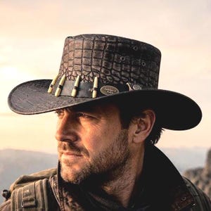 Puede incluir: Un sombrero marrón oscuro con un patrón texturizado de piel de cocodrilo, que presenta una banda negra adornada con cinco detalles decorativos de color claro en forma de diente y una pequeña insignia metálica ovalada con el texto Outback Hats.