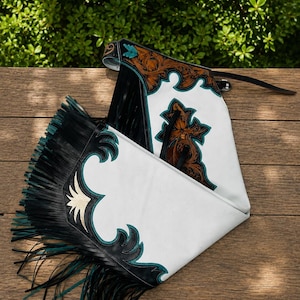 May include: White leather chaps featuring intricate brown floral tooling, black leather overlays, and teal accents. The garment includes long black and teal fringe along the sides and a black leather belt with a buckle closure at the top.