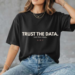 Może przedstawiać: Czarny t-shirt z krótkim rękawem z białym napisem "TRUST THE DATA." pogrubioną czcionką wielkimi literami, po którym następuje mniejszy tekst "not the vibes." oraz litery "A · B · C" wyśrodkowane na klatce piersiowej odzieży.
