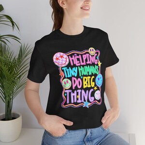 Può includere: Una t-shirt nera a maniche corte con una grafica colorata che riporta il testo "HELPING TINY HUMANS DO BIG THINGS" in rosa, turchese e giallo. Il design è circondato da un bordo viola ondulato, piccole stelle e varie faccine sorridenti.