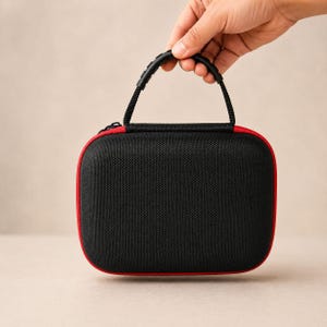 May include: A black carrying case with a red trim and a black handle. The case is rectangular with rounded corners and a zipper closure. The handle is made of a textured, black material.