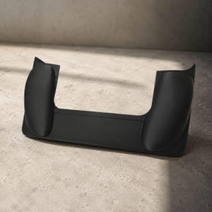 Puede incluir: Un dispositivo negro mate en forma de U con dos lados curvos y elevados y una base plana. El dispositivo está hecho de un material liso y se asienta sobre una superficie de tonos neutros. El diseño sugiere un soporte para un dispositivo electrónico.