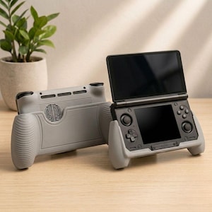 May include: Two gray handheld gaming consoles with black screens and buttons are displayed on a wooden surface. One console is open, showing the screen, while the other is closed. The consoles have gray grips for comfortable handling.