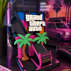 May include: A black, pink, and blue headset stand featuring the Grand Theft Auto V logo in white text. The stand includes two green palm trees, a black base with pink accents, and a small sign that reads WANTED, Jason Deved, and six yellow stars.