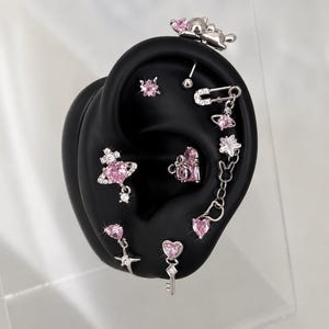 May include: A collection of silver-toned earrings with pink gemstone accents displayed on a black ear-shaped form. The earrings feature various designs, including stars, hearts, keys, and floral shapes. A small cat-shaped earring is also visible.