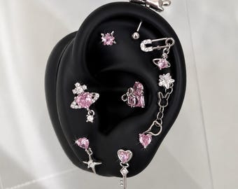 May include: An assortment of silver earrings with pink gemstones, presented on a black ear-shaped display. The earrings showcase diverse designs, such as stars, hearts, keys, and floral motifs. A small cat-shaped earring is also included.
