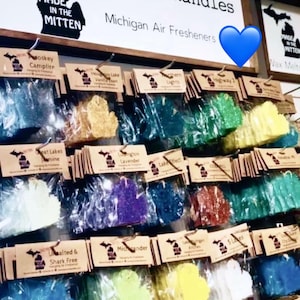 Op de afbeelding: Een display van Michigan-vormige luchtverfrissers in diverse kleuren, hangend aan een muur. Het merk "Kymora Kandles" is zichtbaar, samen met de slogan "Made in the Mitten". Elke luchtverfrisser is verpakt en gelabeld met een geurnaam.