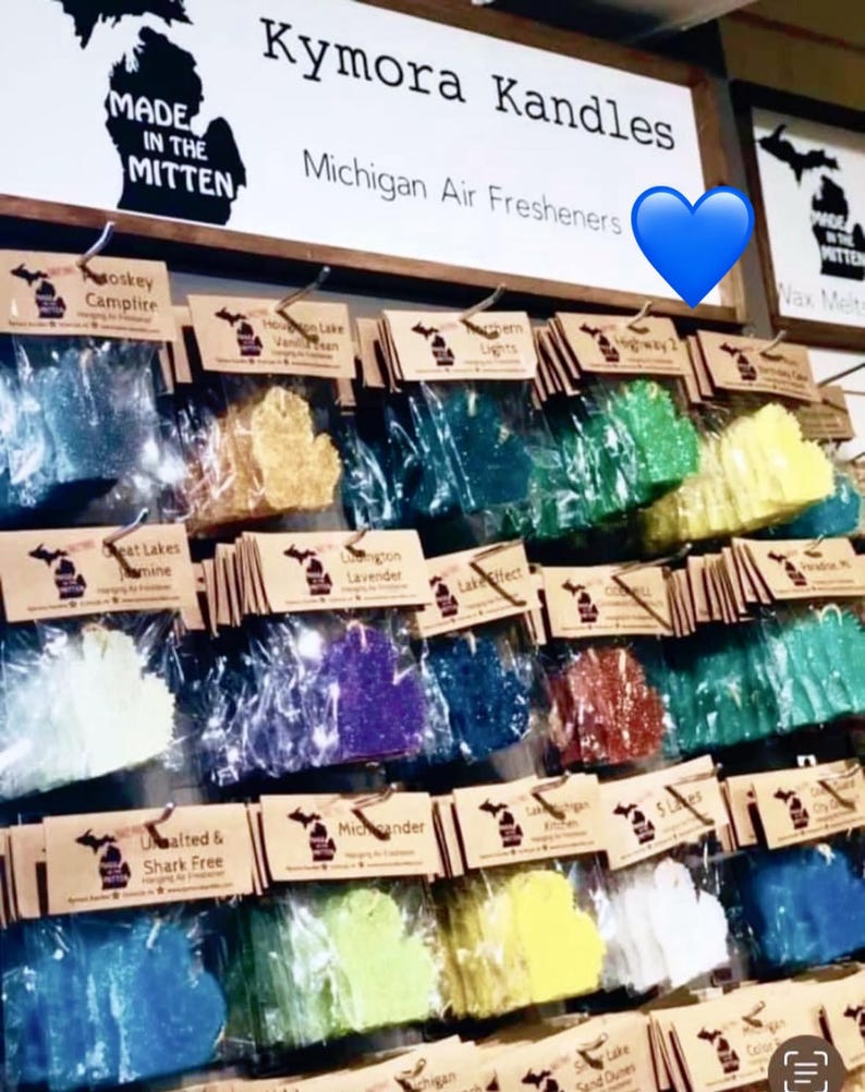 Op de afbeelding: Een display van Michigan-vormige luchtverfrissers in diverse kleuren, hangend aan een muur. Het merk "Kymora Kandles" is zichtbaar, samen met de slogan "Made in the Mitten". Elke luchtverfrisser is verpakt en gelabeld met een geurnaam.