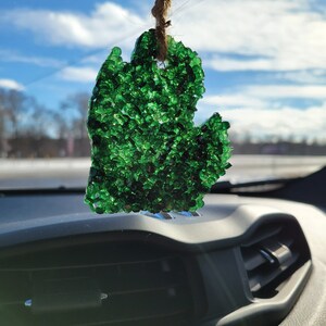 Op de afbeelding: Een groene, Michigan-vormige auto-luchtverfrisser hangt aan een touwtje. De luchtverfrisser is gevuld met kleine, glanzende groene kralen. De achtergrond toont een heldere, zonnige hemel met een paar wolken.
