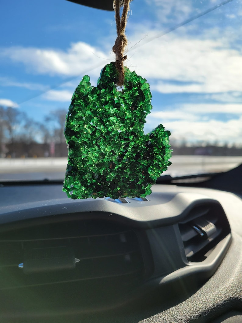 Op de afbeelding: Een groene, Michigan-vormige auto-luchtverfrisser hangt aan een touwtje. De luchtverfrisser is gevuld met kleine, glanzende groene kralen. De achtergrond toont een heldere, zonnige hemel met een paar wolken.