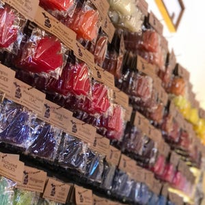Op de afbeelding: Een display van kleurrijke, bladvormige zepen in doorzichtige verpakkingen, elk met een bruin label. De zepen zijn in verschillende kleuren, waaronder rood, oranje, blauw en groen. De labels hebben tekst zoals "Michigan Color Run" en "Apple Orchard".