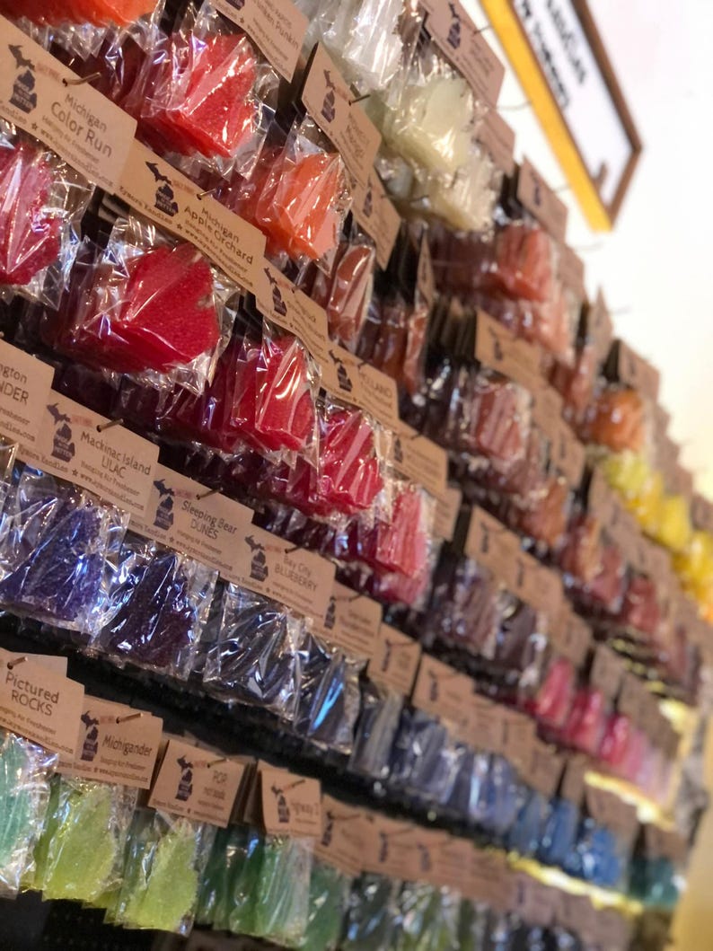 Op de afbeelding: Een display van kleurrijke, bladvormige zepen in doorzichtige verpakkingen, elk met een bruin label. De zepen zijn in verschillende kleuren, waaronder rood, oranje, blauw en groen. De labels hebben tekst zoals "Michigan Color Run" en "Apple Orchard".