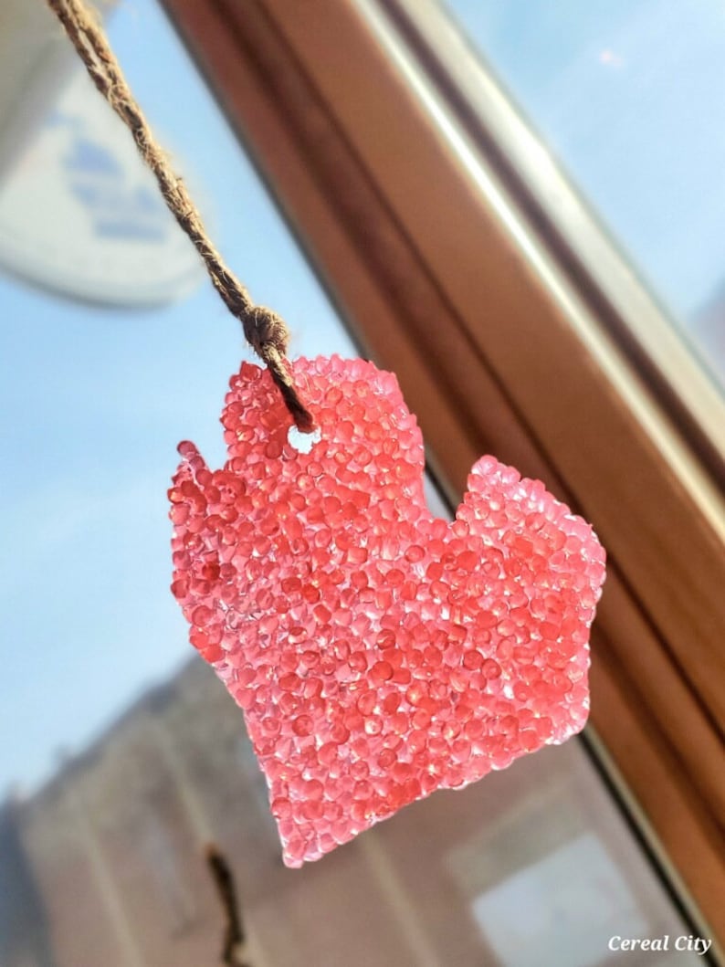Op de afbeelding: Een hartvormige luchtverfrisser, gevuld met kleine, rode kralen, hangt aan een bruin touwtje. De luchtverfrisser hangt voor een raam, met een vage achtergrond van een blauwe lucht en een gebouw.