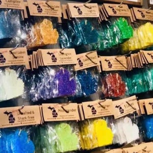 Op de afbeelding: Een display van luchtverfrissers in de vorm van de staat Michigan, in verschillende kleuren. Elk is verpakt in een doorzichtige plastic zak en bevestigd aan een bruin papieren label met tekst. De geuren zijn onder meer Campfire en Houghton Lake Vanilla Bean.